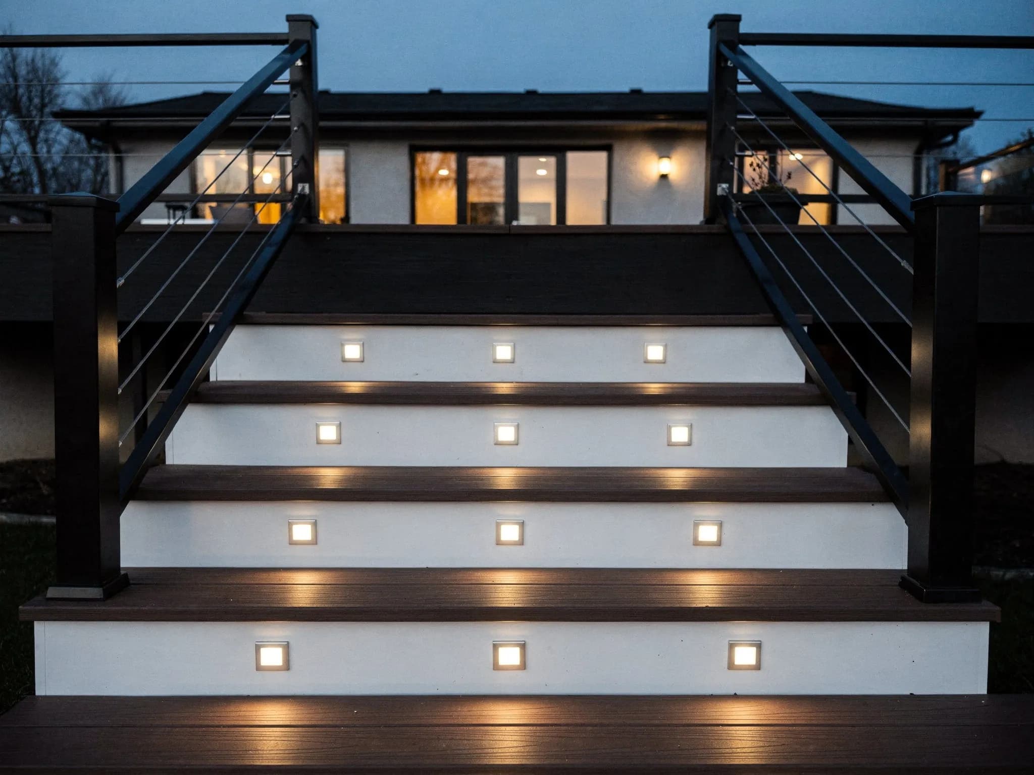 Warm recessed riser lights illuminate wide, dark composite stair treads at dusk, making each step easier to see.